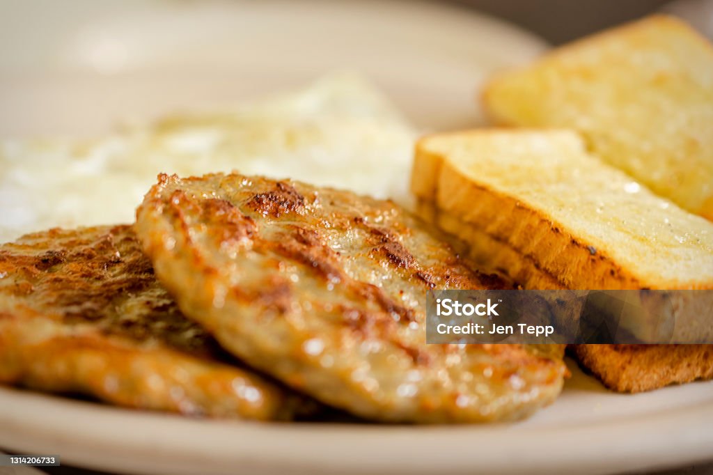 A dish at a breakfast diner including 2 pork sausage patties, two pieces of white toast, and 2 over easy eggs blurred in the background. A dish at a breakfast diner including 2 pork sausage patties, two pieces of white toast, and 2 over easy eggs blurred in the background.  White plate. Sausage Stock Photo
