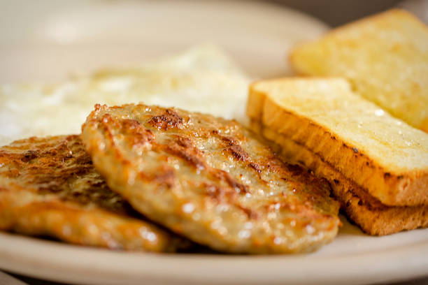 A dish at a breakfast diner including 2 pork sausage patties, two pieces of white toast, and 2 over easy eggs blurred in the background. A dish at a breakfast diner including 2 pork sausage patties, two pieces of white toast, and 2 over easy eggs blurred in the background.  White plate. Sausage stock pictures, royalty-free photos & images