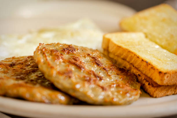 A dish at a breakfast diner including 2 pork sausage patties, two pieces of white toast, and 2 over easy eggs blurred in the background. A dish at a breakfast diner including 2 pork sausage patties, two pieces of white toast, and 2 over easy eggs blurred in the background.  White plate. sausage stock pictures, royalty-free photos & images