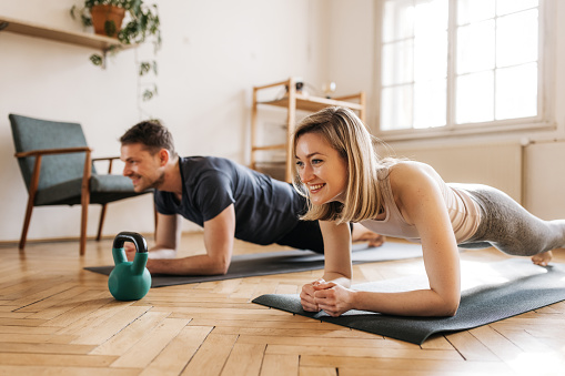 A happy fit couple is finishing their workout in a plank pose. They have big smiles on their faces and are looking forward to successfully completing an intense workout. They are dressed in comfortable sportswear and have matching yoga mats. Luscious light is coming through the window in the back. Horizontal photo.