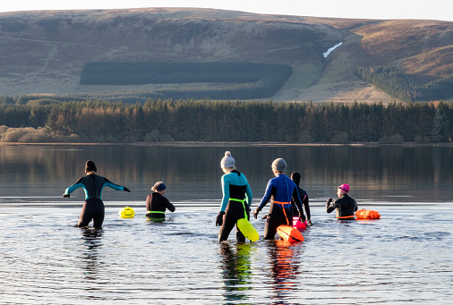 A group of women friends gathering together at the start of the day to wild swim in a Scottish freshwater reservoir. They are wearing wetsuits and warm hats to protect themselves from the cold water. They are wearing brightly coloured inflatable swimming safety belts attached round their waists. The friends stay close together for safety and companionship as they exercise. It is a fun activity that helps with mental health, wellbeing and is enjoyable time out before returning to their families, work and the day ahead.