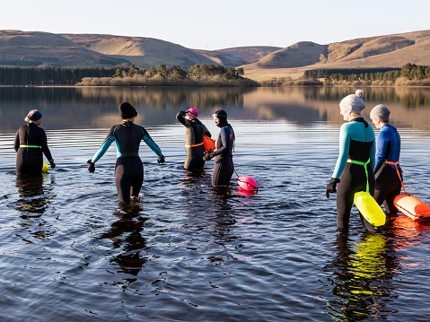 A group of women friends gathering together at the start of the day to wild swim in a Scottish freshwater reservoir, with hills surrounding the water. They are wearing wetsuits and warm hats to protect themselves from the cold water. They are wearing brightly coloured inflatable swimming safety belts attached round their waists. The friends stay close together for safety and companionship as they exercise. It is a fun activity that helps with mental health, wellbeing and is enjoyable time out before returning to their families, work and the day ahead.