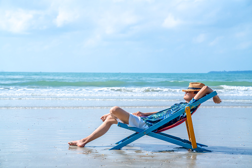 Asian man in casual clothing resting on beach chair at tropical beach. Happy guy sunbathing or nap on sunbed by the sea in sunny day.  Young man relax and enjoy outdoor activity lifestyle on summer holiday vacation