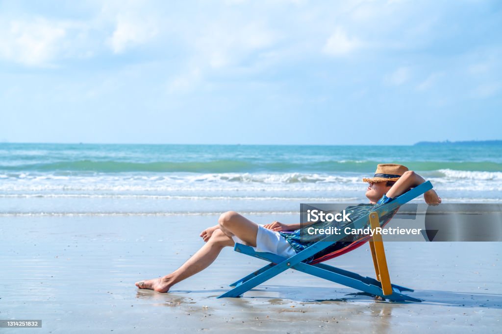 Asian man resting on beach chair on the beach at summer sunny day. Asian man in casual clothing resting on beach chair at tropical beach. Happy guy sunbathing or nap on sunbed by the sea in sunny day.  Young man relax and enjoy outdoor activity lifestyle on summer holiday vacation Relaxation Stock Photo