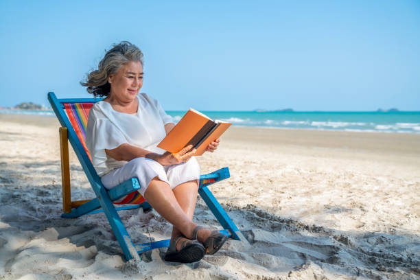 asian senior woman sitting on beach chair with reading a book on the beach - reading outside imagens e fotografias de stock