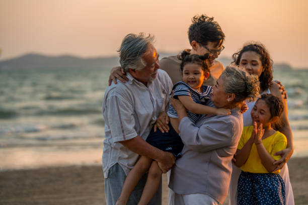 famiglia asiatica multi-generazione che si tiene per mano e cammina insieme sulla spiaggia al tramonto estivo - holding hands couple senior couple togetherness foto e immagini stock