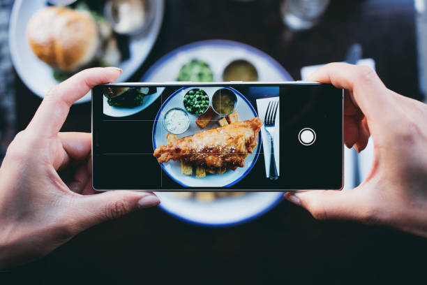 mujer fotografiando pescado y patatas fritas en un restaurante con un smartphone - influencer fotos fotografías e imágenes de stock