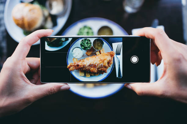 Woman photographing fish and chips in a restaurant with a smartphone Woman photographing fish and chips in a restaurant with a smartphone. english cuisine stock pictures, royalty-free photos & images