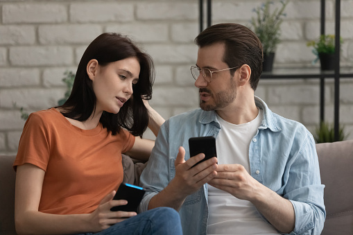 Young family couple sit on couch hold smartphones discuss work of new web app share information between gadgets. Diverse friends man woman users of modern phones talk send receive get photo video file