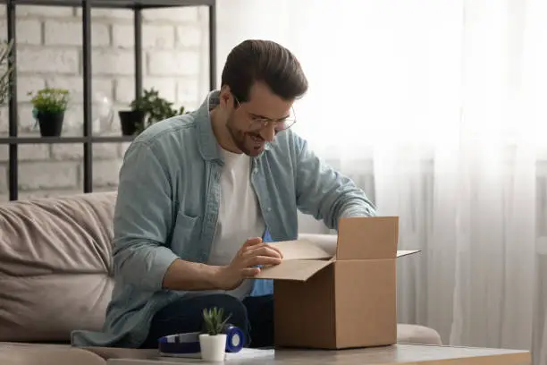 Photo of Smiling young guy opening package with consumer goods with pleasure