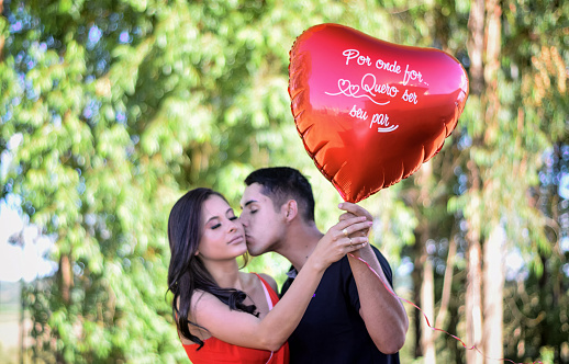 Portrait of a romantic couple, at Valentine’s Day in a forest, in Minas Gerais state, Brazil. The boyfriend is holding a heart balloon saying “Wherever you go, I always want to be your mate” translated from Portuguese, and he is also kissing her cheek. Behind them there are trees from a forest defocussed.