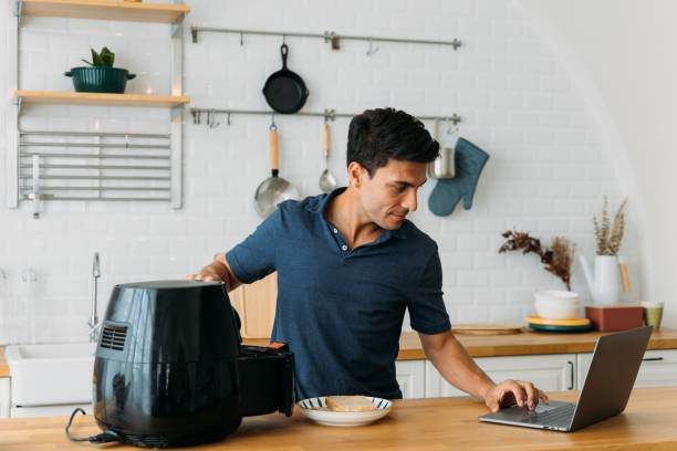 Man using air fryer and laptop in kitchen stock photo