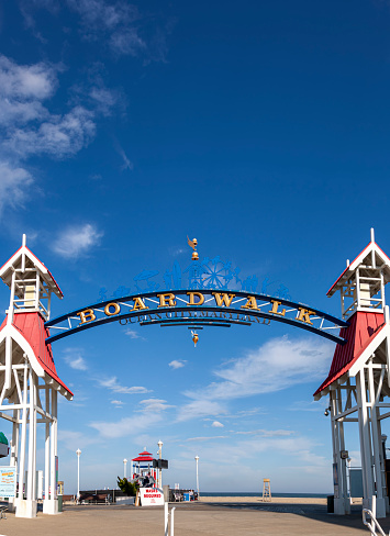 Image  of the entrance of famous board walk of Ocean City, MD by the beach where people are walking on a sunny afternoon. A popular tourist spot on east coast of USA. A wooden arch is erected to mark the spot.