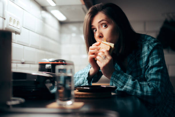 mujer hambrienta comiendo un sándwich por la noche en la cocina - comer demasiado fotografías e imágenes de stock