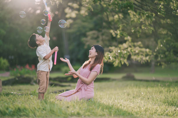 madre china asiática jugando con su hijo con varita de burbujas en el parque público en la luz del sol retroiluminada por la mañana - park child asia lifestyles fotografías e imágenes de stock