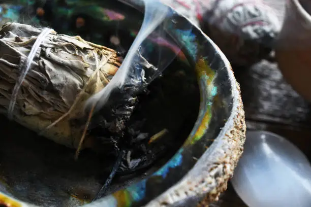 A close up image of a smoldering white sage smudge bundle and healing crystal on a dark wooden table.