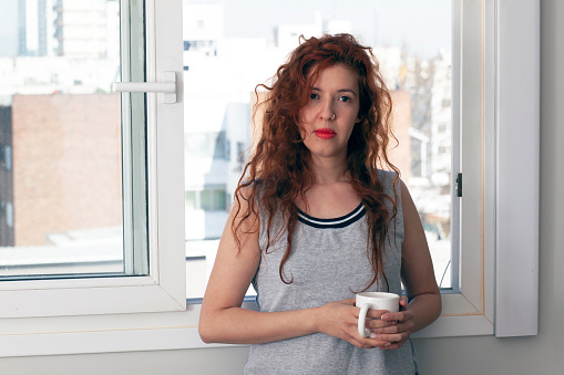 Portrait of young red hair woman holding mug next to open window