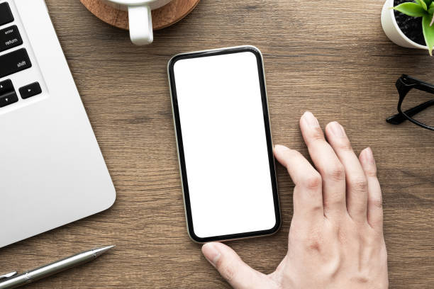 istock Hand holding smartphone with white blank mock up screen over wood office desk table with laptop computer, cup of coffee and supplies. Top view image. 1314289457
