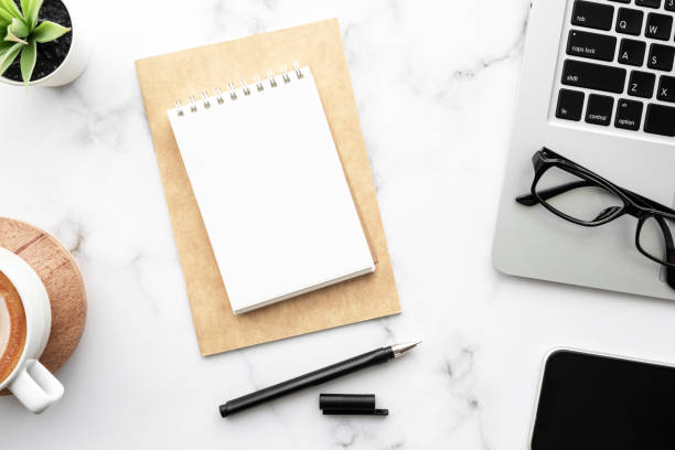 istock Blank notebook with pen are on top of white marble office desk table with laptop computer, cup of coffee and supplies. Top view, flat lay. 1314289528