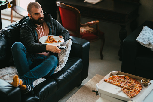 Young casually dressed bearded man sitting at the couch, eating take-out pizza, holding a tv remote and watching tv