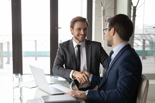 Smiling businessman shaking client hand, closing successful deal, sitting at table with laptop in office, satisfied hr manager hiring new employee, business partners handshaking at meeting