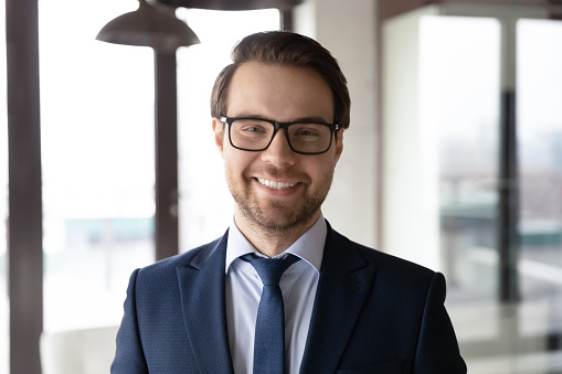 Head shot portrait smiling confident businessman wearing glasses and suit standing in modern office, executive manager company owner employee looking at camera, mentor coach posing for photo