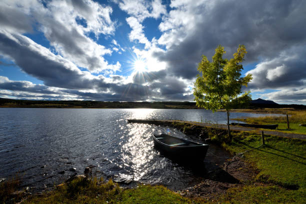 The Loch is in the foreground of the photo. The boat is moored to the bank, and a tree is on its left. The sun is low and there are clouds in the sky giving a moody appearance to the photo.\n\nThe photo was taken in north-west Scotland in October 2020.
