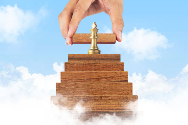Business growth, business success or career path success concept. A king chess is on the top of wooden stacked staircase in cloud scene with a hand is putting the next wooden piece. crecimiento empresarial, éxito empresarial o concepto de éxito de trayectoria profesional. un ajedrez rey está en la parte superior de la escalera apilada de madera en la escena de nubes con una mano está poniendo la siguiente pieza de madera.