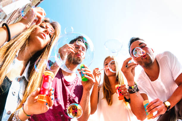 Young friends having fun playing with soap bubbles at spring break time - Friendship concept and genuine life style with happy millenial people together outdoors - Warm bright filter