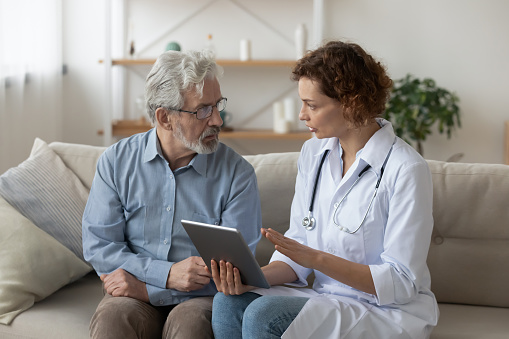 Skilled professional physician doctor prescribing illness treatment, showing health test results on digital computer tablet to concentrated older retired patient at home, sitting together on sofa.