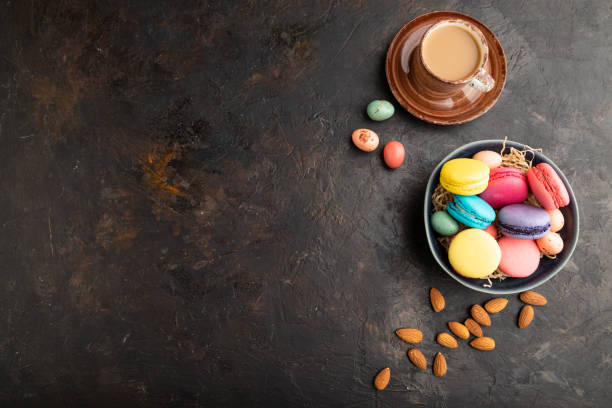 istock Multicolored macaroons and chocolate eggs in ceramic bowl, cup of coffee on black concrete background. top view, flat lay, copy space, still life. Breakfast, morning, concept. 1314334543