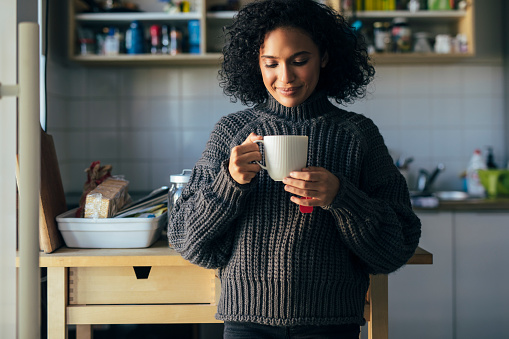 Simple pleasures at home: a young mixed race woman enjoying a cup of hot tea on a winter morning, looking down while facing camera