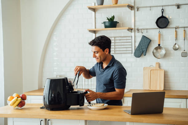 Man using air fryer and laptop in kitchen Handsome young caucasian man looking at instructions online on laptop on how to use modern air fryer to cook healthy food without using oil at home in kitchen deep fryer stock pictures, royalty-free photos & images