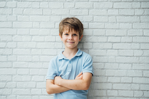 Portrait of happy young caucasian boy in casual outfit with arms crossed isolated over white bricks background smiling and looking at camera