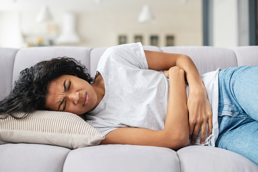 Sad young African American woman wearing casual clothes suffering from menstrual pain, feeling sick to her stomach, holding belly, having abdominal cramps during period and lying down on bed at home