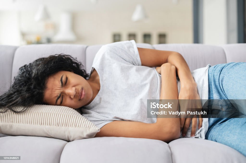 African American woman suffering from cramps Sad young African American woman wearing casual clothes suffering from menstrual pain, feeling sick to her stomach, holding belly, having abdominal cramps during period and lying down on bed at home Irritable Bowel Syndrome Stock Photo