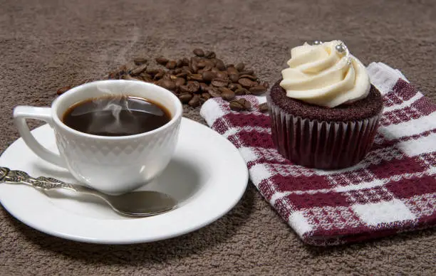 Steaming white coffee cup, homemade cupcake and coffee beans on brown fabric background, homemade cake with cream