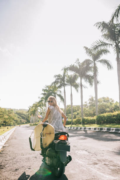 Young woman riding to the beach with motor scooter, to go for a surf.