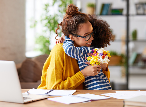 Little child son congratulating mom with Mothers day or birthday, giving her fresh flower bouquet. Happy mixed race woman mother embracing with child while working at home. Family holidays concept