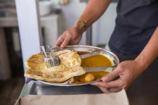 Close-up shot of an unrecognizable male waiter serving Malaysian roti canai with curry sauce. The food is served on a stainless steel compartment plate, with eating utensil above.