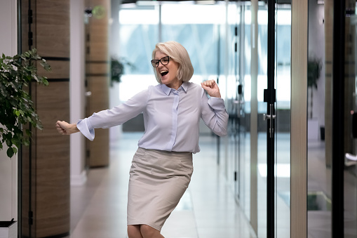 Happy mature Caucasian woman worker dance have fun in office, enjoy Friday time at workplace. Overjoyed middle-aged businesswoman or employee celebrate work achievement or job success.