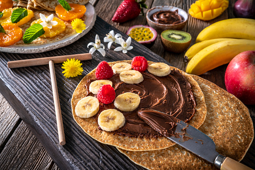 Banana and cocoa cream Crepes arrangement with fruits, berries, cocoa cream and varied vegetarian food still life on rustic wood