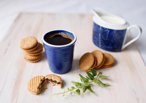 Istanbul, Turkey-April 25, 2021: A blue ceramic coffee mug with drip coffee, a creamer, green leaves and biscuits on a beige light brown wooden background. Shot with Canon EOS R5.