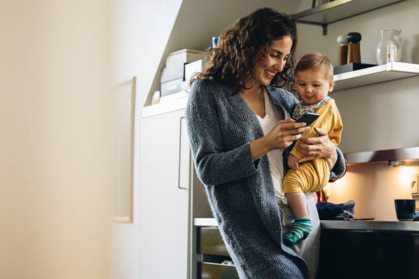 uśmiechnięta kobieta z dzieckiem sms-y na telefon w domu - baby carrier obrazy zdjęcia i obrazy z banku zdjęć