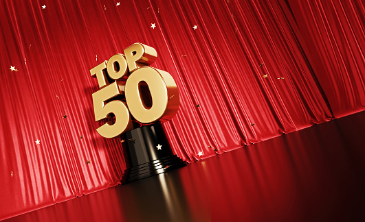 Gold colored confetti falling onto a gold top 50 award sitting before red stage curtain. Horizontal composition with copy space. Low angle view. Top 50 concept.
