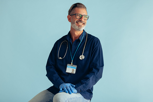 Portrait of a happy mature male doctor on blue background. General practitioner with stethoscope looking at camera and smiling.
