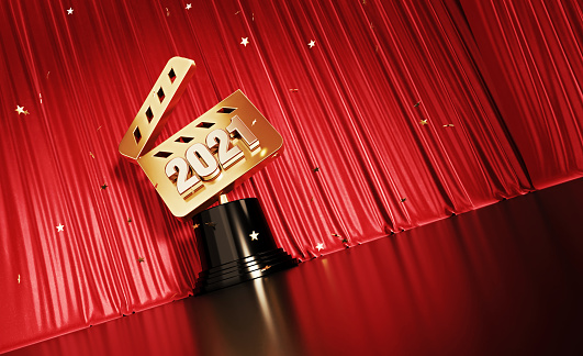 Gold colored confetti falling onto a 2021 written gold film slate award sitting before red stage curtain. Horizontal composition with copy space. Low angle view.