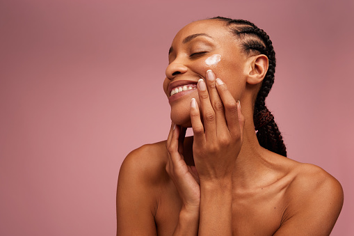 Beautiful woman eyes closed while applying moisturizer to her face. Woman applying face cream against pink background.
