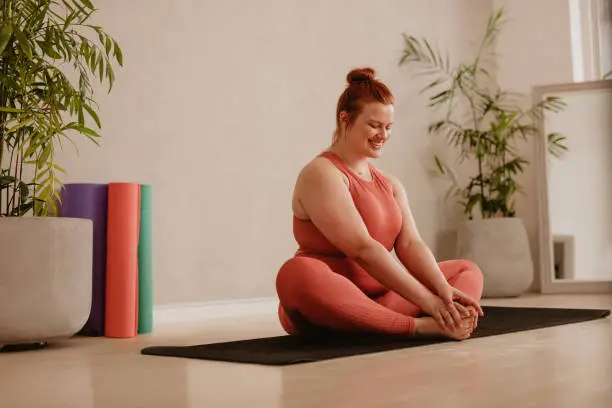Plus size woman doing yoga stretching workout at fitness studio. Female wearing sportswear sitting on exercise mat and doing stretching workout at gym.