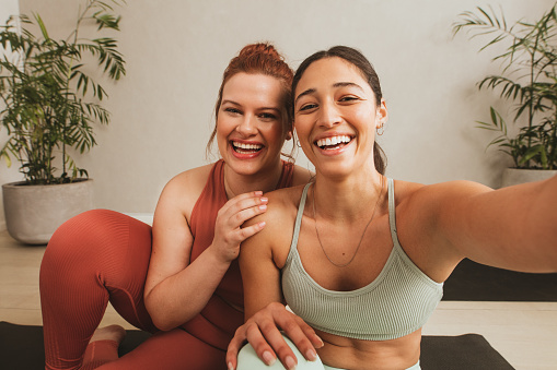 Cheerful female friends taking selife at yoga gym. Women in sports wear taking a break from workout and talking selfie at fitness studio.